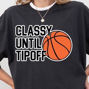 May include: A black crewneck sweatshirt featuring the white text "CLASSY UNTIL TIPOFF" stacked vertically next to a graphic of an orange basketball with black lines. The design is centered on the chest of the garment.