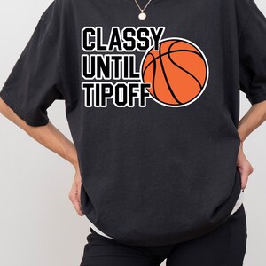Op de afbeelding: Een zwart T-shirt met korte mouwen en de witte tekst "CLASSY UNTIL TIPOFF" verticaal gestapeld naast een afbeelding van een oranje basketbal met zwarte lijnen. Het shirt heeft een ronde hals en een relaxte pasvorm van zwarte stof.
