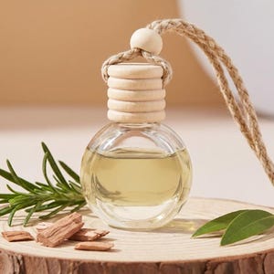 May include: A small, round, clear glass bottle filled with a pale yellow liquid, topped with a ribbed light-colored wooden cap. A thick, braided jute rope is attached to the top for hanging. A sprig of green rosemary and small wood chips sit nearby.