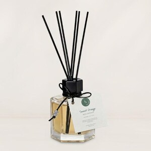 May include: A clear glass hexagonal reed diffuser bottle filled with light amber liquid. It features a black cap, several black reed sticks, and a black string tied around the neck with a silver bead. A white tag reads Sweet Orange REED DIFFUSER.