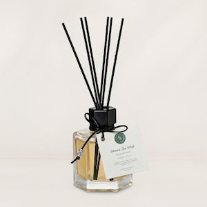 May include: A clear hexagonal glass reed diffuser bottle filled with light amber liquid. It features a black cap, six black reeds, and a black string tied around the neck with a silver bead. A white tag reads Green Tea Mint REED DIFFUSER.