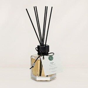 May include: A hexagonal glass reed diffuser filled with light amber liquid, featuring a black cap and six black reeds. A black cord with a silver bead secures a white rectangular tag printed with the text Hippie Reed Diffuser and other details.