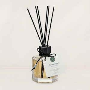 May include: A clear glass hexagonal reed diffuser bottle filled with light amber liquid, topped with a black cap and six black reeds. A black string ties a white rectangular tag to the neck, featuring the text Rosemary Mint Reed Diffuser.