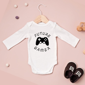 Könnte beinhalten: Weißer Baby-Body mit langen Ärmeln, mit einem schwarzen Controller-Motiv und dem Text "FUTURE GAMER". Der Body hängt an einem Holzbügel. Braune Babyschuhe liegen daneben.