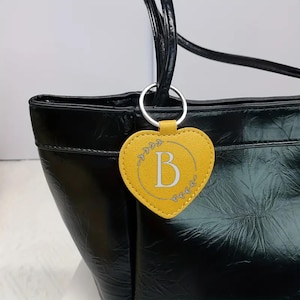May include: A yellow heart-shaped keyring featuring the letter "B" in the centre, with a silver floral design. The keyring is attached to a black tote bag with a silver ring.