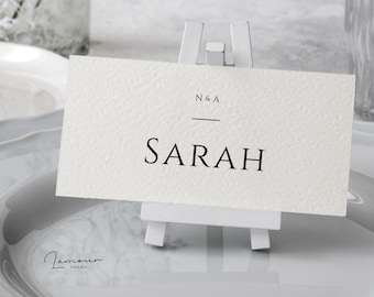 Può includere: Segnaposto bianco con testo nero che recita "N & A" e "SARAH" su un piccolo cavalletto bianco.