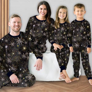 Könnte beinhalten: Eine vierköpfige Familie trägt passende schwarze Pyjamas mit einem goldenen und weißen Feuerwerksmuster. Die Pyjamas haben einen dunkelblauen Besatz. Die Familie sitzt auf einem weißen Ottoman in einem gut beleuchteten Raum.
