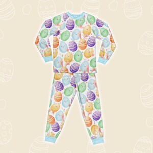 May include: Easter pyjama set with a repeating pattern of cartoon Easter eggs in various colours, some with faces and bunny ears. The long-sleeved top and trousers have light blue trim and are set against a cream background with egg outlines.