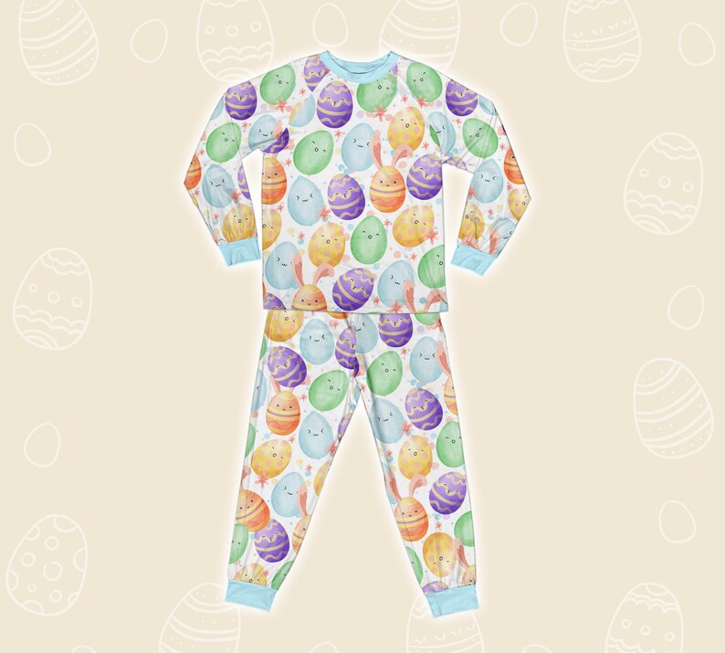 May include: Easter pyjama set with a repeating pattern of cartoon Easter eggs in various colours, some with faces and bunny ears. The long-sleeved top and trousers have light blue trim and are set against a cream background with egg outlines.