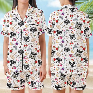 K&ouml;nnte beinhalten: Wei&szlig;es Pyjama-Set mit einem skurrilen Muster aus schwarzen Drachen, Rosen, B&uuml;chern, Monden und anderen Fantasy-Elementen. Das kurz&auml;rmelige Oberteil und die Shorts haben schwarze Paspeln.
