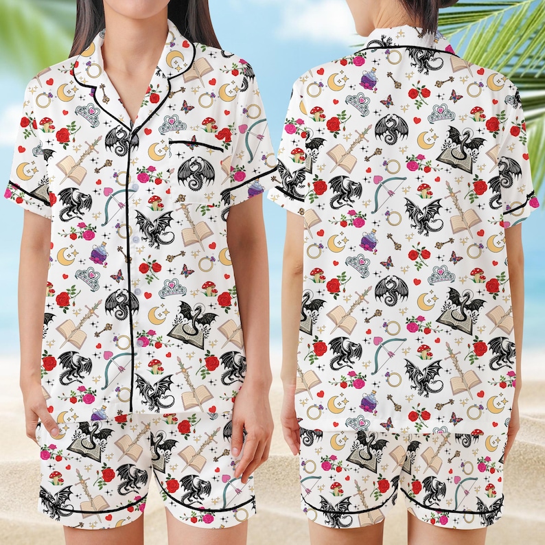 K&ouml;nnte beinhalten: Wei&szlig;es Pyjama-Set mit einem skurrilen Muster aus schwarzen Drachen, Rosen, B&uuml;chern, Monden und anderen Fantasy-Elementen. Das kurz&auml;rmelige Oberteil und die Shorts haben schwarze Paspeln.