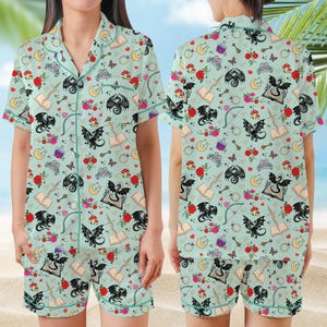 K&ouml;nnte beinhalten: Hellblaues Pyjama-Set mit einem sich wiederholenden Muster aus schwarzen Drachen, Rosen, B&uuml;chern, Monden und anderen Fantasy-Elementen. Das kurz&auml;rmlige Oberteil und die Shorts haben hellblaue Paspeln. Der Stoff ist weich und leicht.