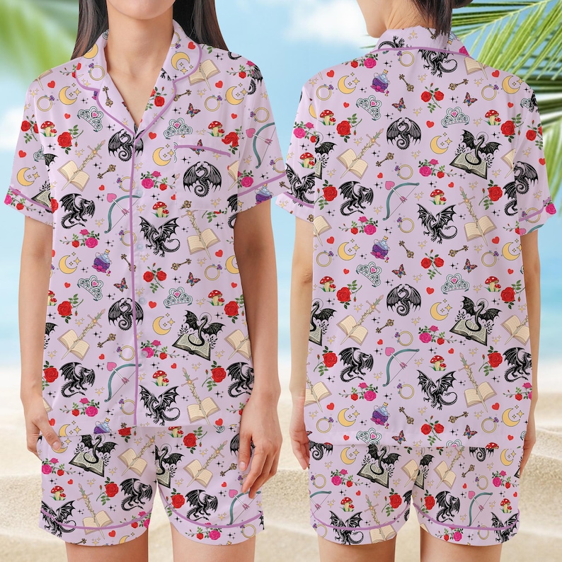 K&ouml;nnte beinhalten: Helllila Pyjama-Set mit einem sich wiederholenden Muster aus schwarzen Drachen, Rosen, B&uuml;chern, Monden und anderen Fantasy-Elementen. Das kurz&auml;rmelige Oberteil und die Shorts haben lila Paspeln.