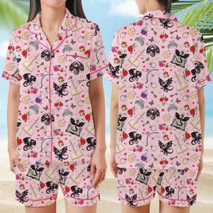 K&ouml;nnte beinhalten: Rosa Pyjama-Set mit einem skurrilen Muster aus schwarzen Drachen, Rosen, Pilzen, Monden und B&uuml;chern. Das kurz&auml;rmlige Oberteil und die Shorts haben rote Paspeln.