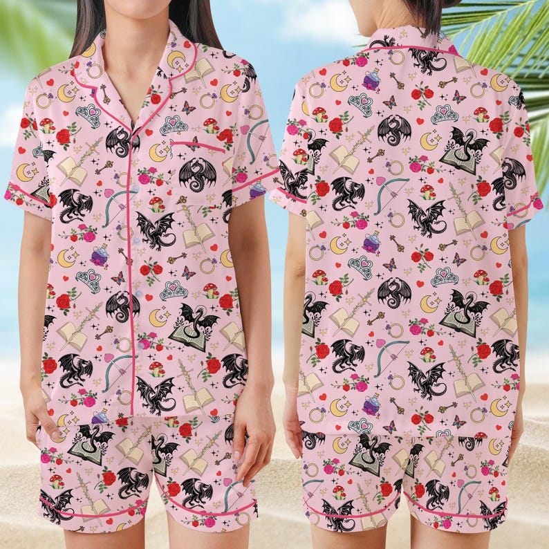 K&ouml;nnte beinhalten: Rosa Pyjama-Set mit einem skurrilen Muster aus schwarzen Drachen, Rosen, Pilzen, Monden und B&uuml;chern. Das kurz&auml;rmlige Oberteil und die Shorts haben rote Paspeln.