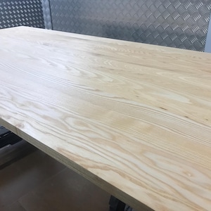 May include: A large, unfinished wooden table with a light, natural wood grain. The rectangular table is made of solid wood. The wood grain is visible across the surface. The table is supported by a black metal frame. The table's dimensions are in centimetres.