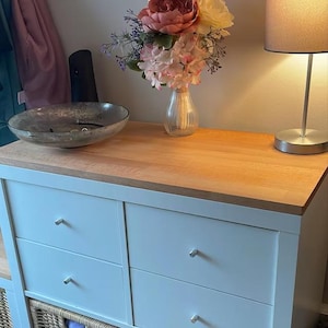 May include: A white dresser with four drawers, each featuring a small silver knob. The top surface is light-colored wood. On top sits a clear glass vase with pink and white artificial flowers, a shallow gray bowl, and a silver table lamp.