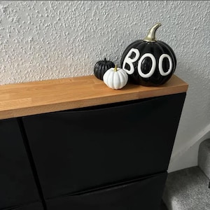 May include: Black shoe storage cabinet with three drawers. The cabinet has a light wood top and is decorated with three Halloween-themed pumpkins, one white and two black, one of which says "BOO" in white letters.