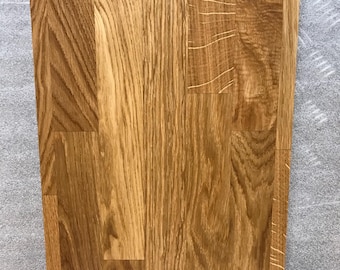Puede incluir: Primer plano de una tabla de cortar rectangular de madera. La tabla es de madera de color marrón claro con un patrón de vetas visible. La madera está dispuesta en secciones verticales y horizontales, creando una superficie texturizada.