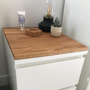 Può includere: Un comodino bianco con un piano in legno chiaro. Una lampada nera con paralume in vetro trasparente e una piccola pianta grassa in vaso sono sulla parte superiore. Un tubetto di lozione alla lavanda e un piccolo blocco di legno sono presenti.