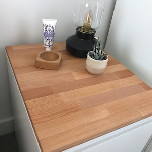 May include: A white nightstand with a light wood top. On the top are a black lamp with a glass shade, a small succulent in a white pot, a wooden bowl, and a tube of hand cream.