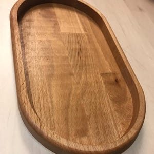 Puede incluir: Una bandeja ovalada de madera natural con un borde elevado. La bandeja está hecha de madera marrón claro y tiene un acabado liso.