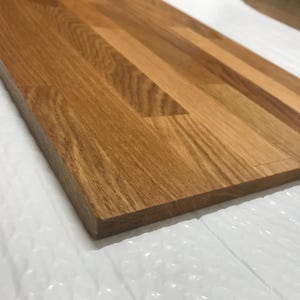 Könnte beinhalten: Nahaufnahme einer rechteckigen Holzplatte mit natürlicher Maserung. Die Platte hat eine warme braune Farbe und scheint aus mehreren Holzbrettern zusammengesetzt zu sein. Die Kanten sind sauber und die Oberfläche ist glatt.