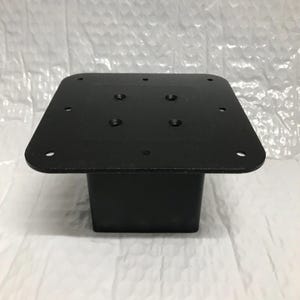May include: Black metal square base with multiple screw holes on the top surface. The base is approximately 15 cm wide and 10 cm tall.