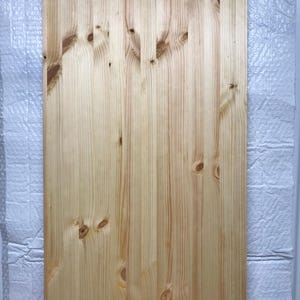 May include: A rectangular, unfinished wooden panel with a light, natural wood tone. The panel features vertical grain patterns and visible knots, showcasing the wood's natural characteristics. The panel is likely for woodworking projects or furniture making.