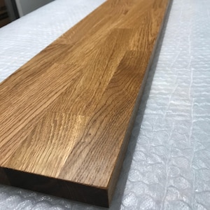 May include: A long, rectangular wooden board featuring a natural light brown oak grain pattern. The surface is smooth and finished with a clear coating, showing visible wood joints where smaller pieces of timber are joined together in a linear fashion.