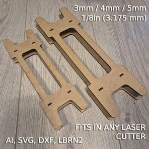 以下が含まれることがあります： 3mm、4mm、または5mm（1/8インチ）厚の素材用のレーザーカットされた木製スタンド2つ。スタンドは、あらゆるレーザーカッターに適合するように設計されています。木には「FITS IN ANY LASER CUTTER」と「AI、SVG、DXF、LBRN2」のテキストが印刷されています。