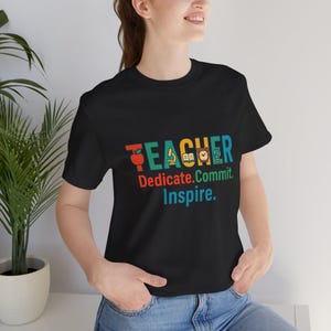 May include: Black t-shirt with the word "TEACHER" in colorful letters, featuring apple, microscope, book, and globe icons. Below, the words "Dedicate. Commit. Inspire." are printed in red, blue, and green.