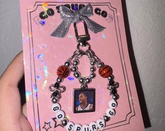 May include: A keychain featuring a silver lobster clasp, a metallic silver bow, and a circular beaded strand. The strand includes orange basketball beads, silver spacer beads, small black bows, and white letter beads spelling GO SPURS GO. A square charm displays a photograph of a basketball player.