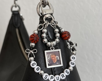 May include: A circular bag charm featuring silver-toned beads, two orange basketball-shaped beads, and small silver bows. A square pendant displays a photograph of a basketball player, while white letter beads spell out GO SPURS GO.