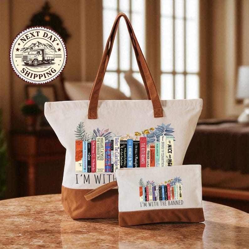 A white tote bag with a printed design of colorful books and the text "I'm with the Banned" printed on it, along with a smaller bag or pouch with the same design.