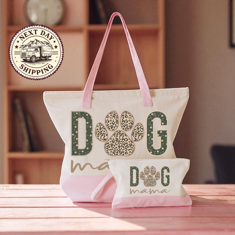 A pink tote bag with a leopard print paw print design and the word "dog" printed on it, along with a smaller bag that says "dog mama" on it. The bags are placed on a wooden surface, and there is a clock visible in the background.