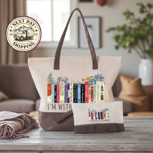 A beige tote bag with a printed design of colorful books and the text "I'm with the Banned" printed on it, along with a smaller bag or pouch with the same design.