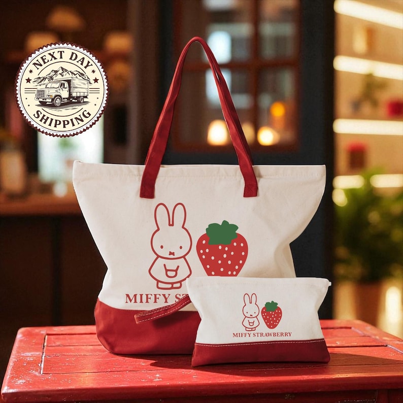 A red and white tote bag with a cartoon rabbit and a strawberry on it, along with a smaller bag featuring a similar design, placed on a red wooden surface.