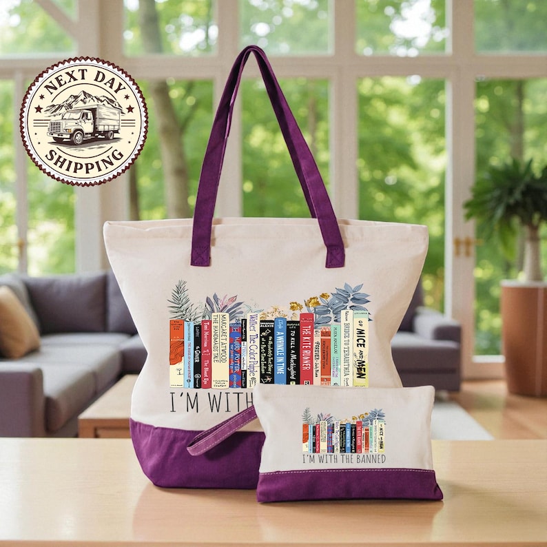 A large tote bag with a printed design of colorful books and the text "I'm with the Banned" printed on it, along with a smaller bag that also has the same design. The bags are placed on a wooden table, and there is a window in the background.