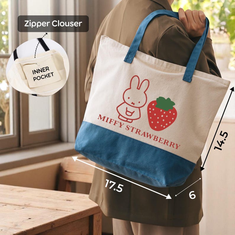 A person is holding a large tote bag with a cartoon rabbit and a strawberry on it. The bag has a zipper closure and a pocket on the front. The bag is described as having a blue and white color scheme.