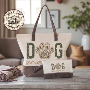 A beige tote bag with a leopard print paw print design and the word "dog" printed on it, along with a smaller bag that says "dog mama" on it. The bag is placed on a wooden surface, and there is a potted plant in the background.
