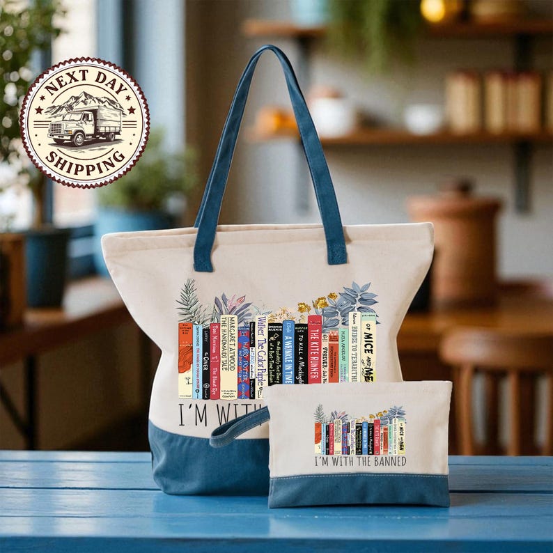 A large tote bag with a printed design of colorful books and the phrase "I'm with the Banned" printed on it, along with a smaller bag below it. The bags are placed on a blue surface, and there is a shipping label visible in the background.