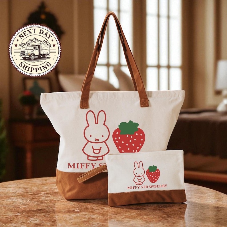 A white tote bag with a cartoon rabbit and a strawberry on it, along with a smaller white bag featuring a cartoon rabbit and a strawberry. The bag is placed on a table, and there is a sign in the background that says "NEXT DAY SHIPPING".
