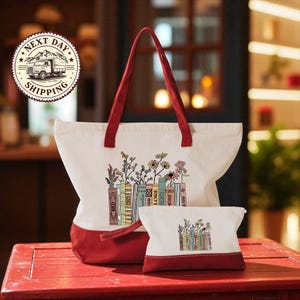 A large white tote bag with a red handle and a smaller white makeup bag with a similar design, both featuring illustrations of books and flowers, are displayed on a red wooden surface.