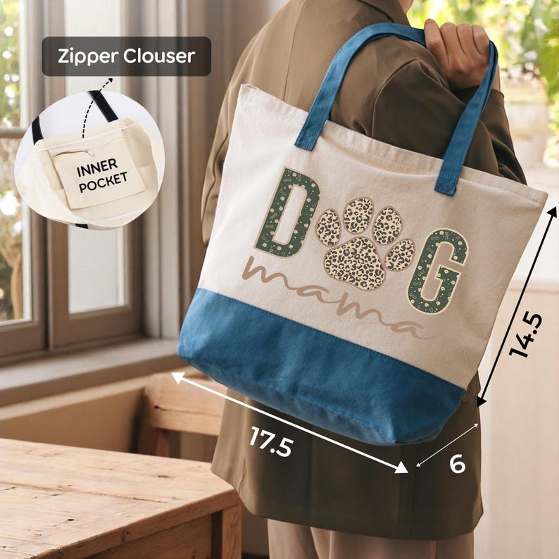 A person is holding a large tote bag with a dog paw print design and the word "dog mama" printed on it. The bag has a zipper closure and a pocket on the front.
