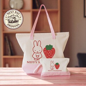 A pink tote bag with a cartoon rabbit and a strawberry on it, along with a smaller bag, is displayed on a wooden surface.
