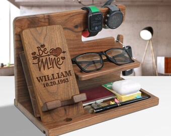 May include: A wooden desk organizer with multiple compartments. It features a phone stand, watch holders, and a tray for accessories. The organizer is engraved with "Be Mine" and a name and date. A pair of glasses and two smartwatches are visible.