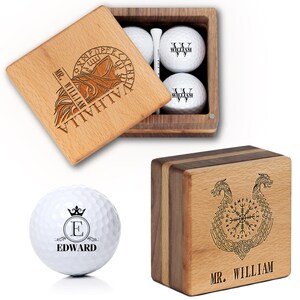 May include: Wooden golf ball gift set. The set includes a wooden box with engraved wolf and text, golf balls with custom initials, and a wooden box with a Viking design. The golf balls are white.