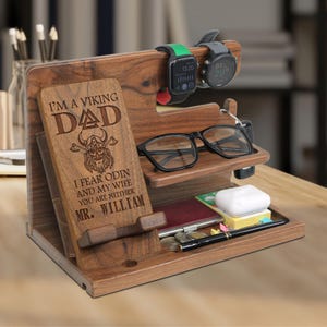 May include: A wooden desk organizer with multiple compartments. It features a slot for a phone, a shelf for glasses, and a section for small items. The organizer has a personalized plaque that reads "I'M A VIKING DAD". Two smartwatches are displayed on top.