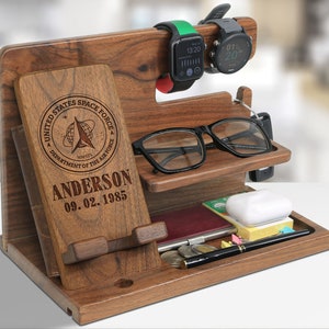 May include: A wooden desk organizer with multiple compartments. It features a phone stand, watch holders, and a tray for accessories. The organizer has the text "UNITED STATES SPACE FORCE" and "ANDERSON 09.02.1985" engraved on it. Two smartwatches and eyeglasses are also visible.